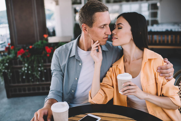 Sensual couple with closed eyes sitting together at table with cups of coffee and man hugging sensual girlfriend leaning closer to kiss - Φωτογραφία, εικόνα