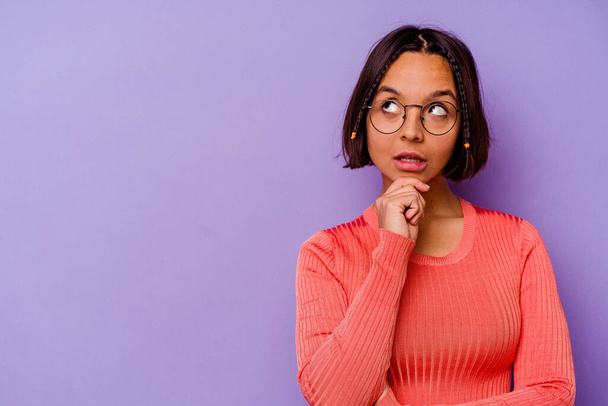 Молодая женщина смешанной расы, изолированная на фиолетовом фоне, смотрит в сторону с сомнительным и скептическим выражением. - Фото, изображение