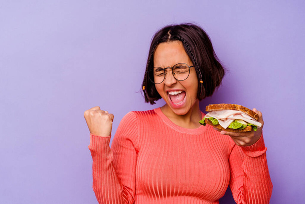 Mladá smíšená závodnice drží sendvič izolovaný na fialovém pozadí zvedající pěst po vítězství, vítěz koncept. - Fotografie, Obrázek
