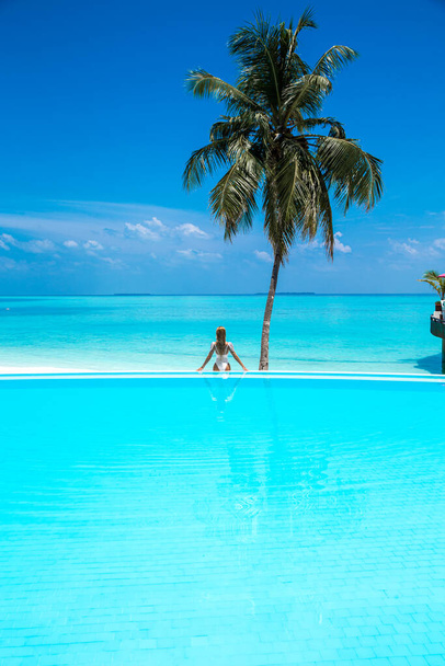 Elegante sexy Frau im weißen Bikini mit dem sonnengebräunten schlanken und wohlgeformten Körper posiert in der Nähe des Swimmingpools auf den Malediven. Perfektes Body-Bikini-Modell im Luxus-Resort auf den Malediven. Luxusreisen. Rückansicht. Rückseite. - Foto, Bild