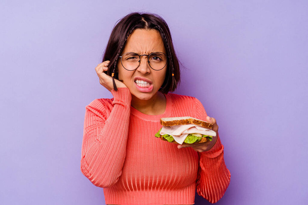 Молода змішана расова жінка тримає сендвіч ізольовано на фіолетовому фоні, покриваючи вуха руками
. - Фото, зображення