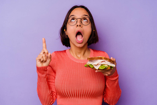 Jeune femme métisse tenant un sandwich isolé sur fond violet pointant à l'envers avec bouche ouverte. - Photo, image