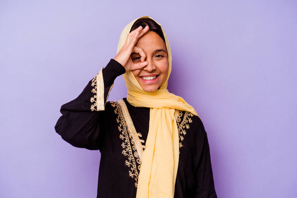 Молода араб, одягнена в типовий арабський костюм, ізольована на пурпуровому фоні, захоплена тим, щоб тримати в порядку жести на око. - Фото, зображення