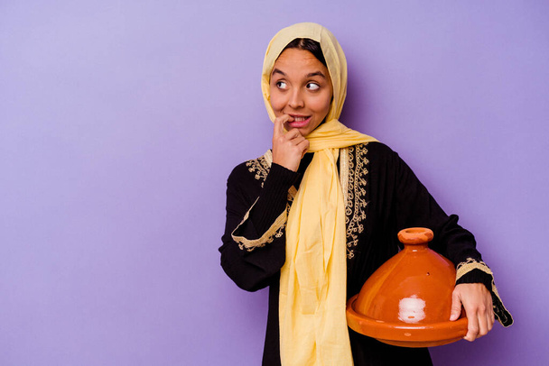 Giovane donna marocchina in possesso di un tajine isolato su sfondo viola rilassato pensando a qualcosa guardando uno spazio copia. - Foto, immagini
