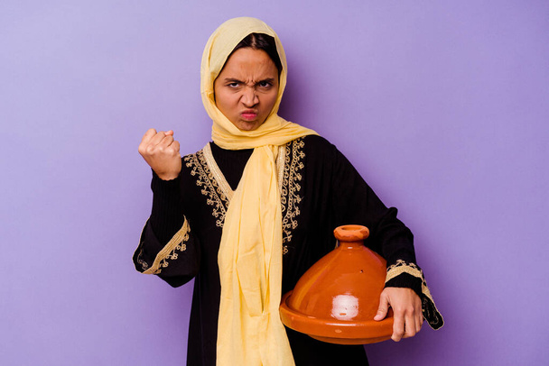 Joven mujer marroquí sosteniendo un tajine aislado sobre fondo púrpura mostrando puño a cámara, expresión facial agresiva. - Foto, imagen