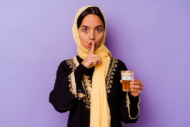 Junge Marokkanerin hält ein Glas Tee isoliert auf violettem Hintergrund und hält ein Geheimnis oder bittet um Schweigen. - Foto, Bild