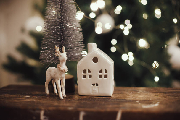 Stylish christmas tree, reindeer toy and house on rustic wood on background of christmas lights. Beautiful little christmas scene. Merry christmas. Magical christmas time. Space for text - Photo, Image
