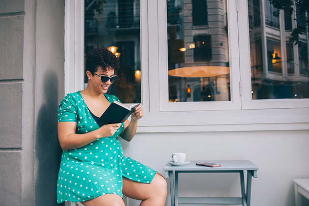 Bright plump ethnic lady in casual clothes and sun glasses reading novel enjoying time sitting at table at outside of cafe - Photo, image