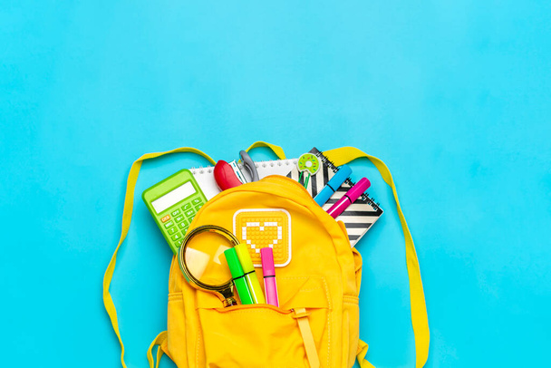 Back to school, education concept. Yellow backpack with school supplies - notebook, pens, ruler, calculator, scissors isolated on blue background. Top view. Copy space Flat lay composition - Photo, Image