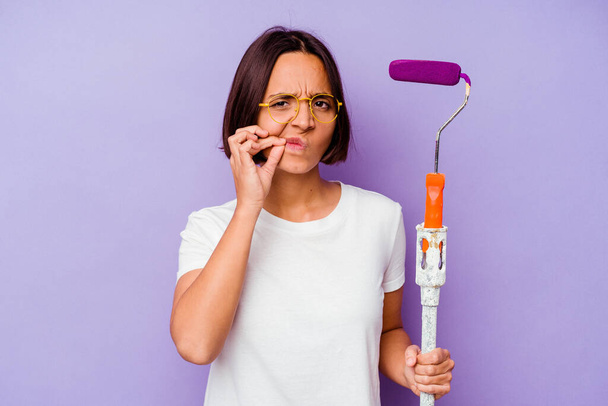 Young painter mixed race woman holding a paint stick isolated on purple background with fingers on lips keeping a secret. - Foto, Bild