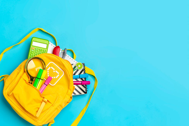 Back to school, education concept. Yellow backpack with school supplies - notebook, pens, ruler, calculator, scissors isolated on blue background. Top view. Copy space Flat lay composition - Zdjęcie, obraz