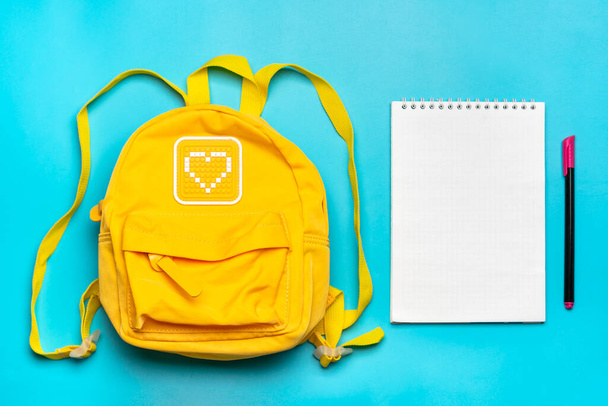 Wracając do szkoły, koncepcja edukacji. Żółty plecak z materiałami szkolnymi - notatnik, długopis odizolowany na niebieskim tle. Widok góry. Przestrzeń kopiowania Kompozycja płaska lay - Zdjęcie, obraz