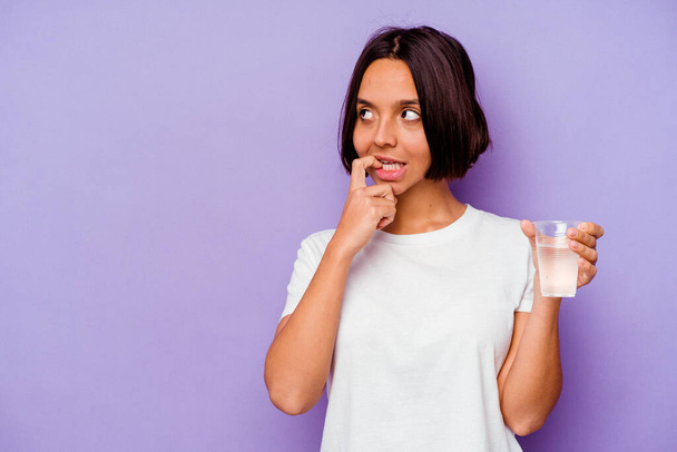 Young mixed race holding a glass of water isolated on purple background relaxed thinking about something looking at a copy space. - Foto, immagini