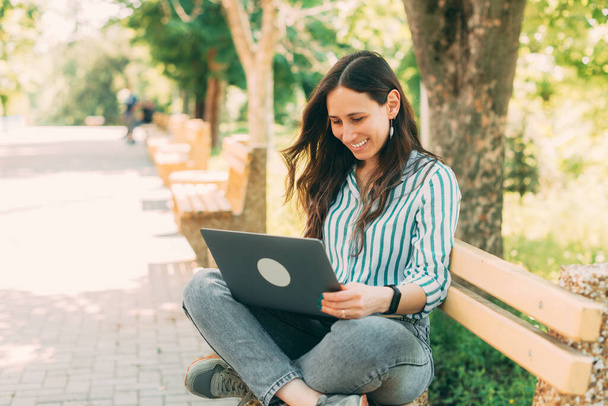 Smiling woman is typing and looking at her laptop on a bench in a park on a beautiful day. - Photo, Image