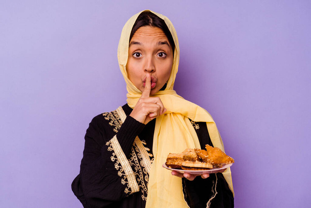 Jonge Marokkaanse vrouw met een Arabisch snoep geïsoleerd op paarse achtergrond houden van een geheim of vragen om stilte. - Foto, afbeelding