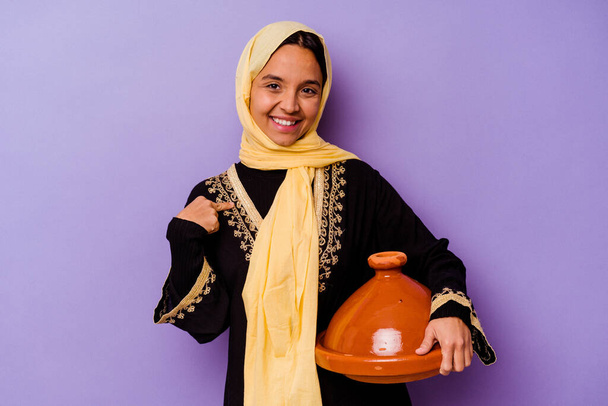 Joven mujer marroquí sosteniendo un tajine aislado sobre fondo púrpura persona señalando a mano a un espacio de copia de la camisa, orgulloso y seguro - Foto, imagen