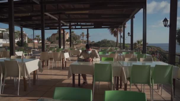 孤独な悲しい仮面の男が座って、アヒオス・ジョージアの空のレストランで自分自身のスマートフォンを使用しています。捨てられたカフェに座っている間、マスクを身に着けている訪問者は注文を待ちます,社会的距離と隔離. - 映像、動画