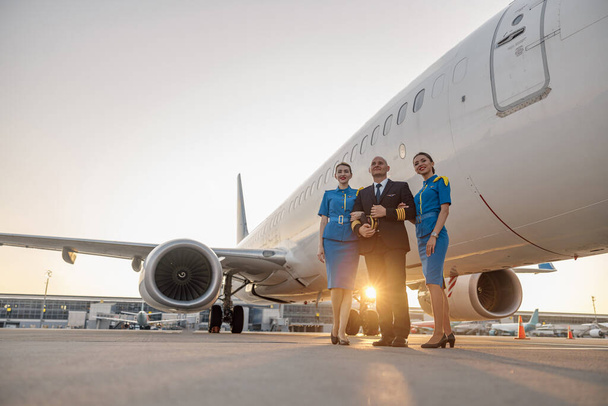 Pełna długość ujęcia samca pilota pozującego razem z dwiema stewardesami w niebieskim mundurze przed samolotem w terminalu o zachodzie słońca - Zdjęcie, obraz