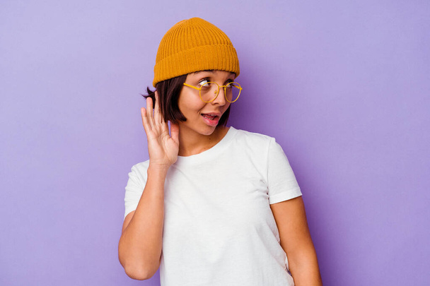 jong gemengd ras vrouw het dragen van een wol cap geïsoleerd op paarse achtergrond proberen om te luisteren naar een roddel. - Foto, afbeelding