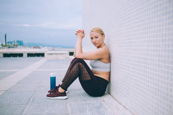 Side view of young female in sportswear looking at camera and sitting behind water bottle while taking break after workout on rooftop - Foto, Imagen