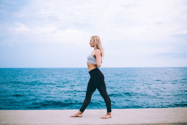 Side view of young flexible focused blond female in black leggings and white top practicing yoga asana on bank of sea - 写真・画像