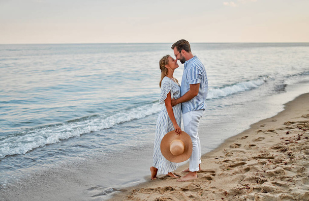 Kochająca młoda para przytula się nad brzegiem morza, ciesząc się sobą i swoimi wakacjami. Kobieta w sukience i mężczyzna w koszuli i białych spodniach spacerują po plaży.. - Zdjęcie, obraz