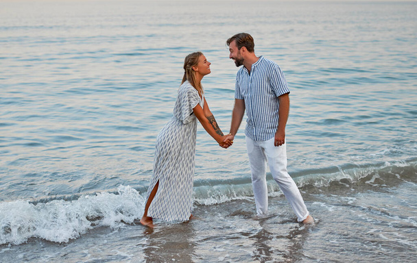Μια ελκυστική γυναίκα με φόρεμα και ένας όμορφος γενειοφόρος άντρας με ριγέ πουκάμισο κρατιούνται χέρι χέρι και ρομαντικά περνούν το χρόνο τους στην παραλία. Ένα ερωτευμένο ζευγάρι σε διακοπές. - Φωτογραφία, εικόνα