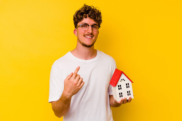 Jovem caucasiano segurando uma casa modelo isolada no fundo amarelo apontando com o dedo para você como se convidando chegar mais perto. - Foto, Imagem