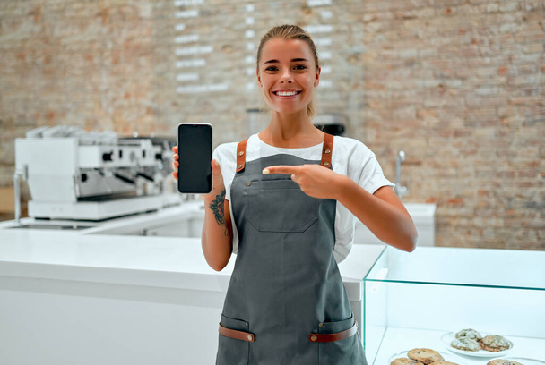 Joven mujer atractiva barista se para en el mostrador en una cafetería y sonríe mientras muestra una pantalla de teléfono inteligente en blanco. - Foto, Imagen