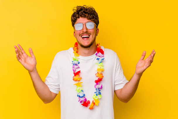 Młody biały mężczyzna ubrany w hawajskie rzeczy na żółtym tle otrzymuje miłą niespodziankę, podniecony i podnoszące ręce. - Zdjęcie, obraz