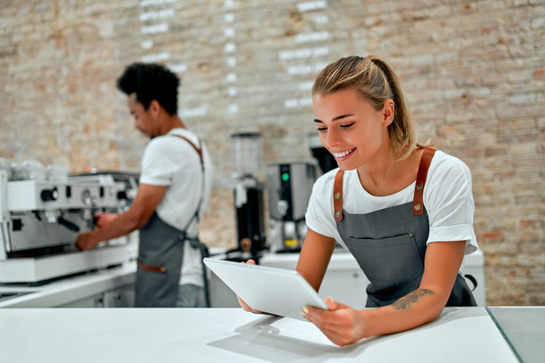 Blanke aantrekkelijke vrouw barista staat met een tablet in haar handen aan de toonbank in een coffeeshop. - Foto, afbeelding