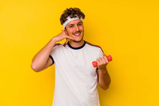 Joven hombre caucásico deportivo sosteniendo una mancuerna aislada sobre un fondo amarillo que muestra un gesto de llamada telefónica móvil con los dedos. - Foto, imagen