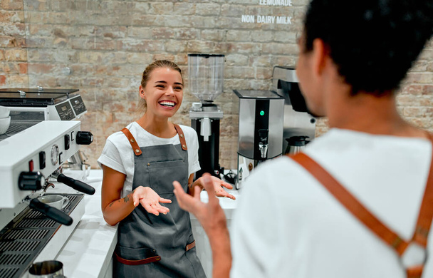 Beautiful caucasian woman and attractive african barista male dressed in apron uniform chatting and laughing in the coffee shop. - Foto, afbeelding