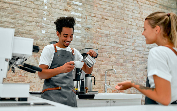Beautiful caucasian woman and attractive african barista male dressed in apron uniform are making coffee, chatting and laughing in the cafeteria. - Photo, Image