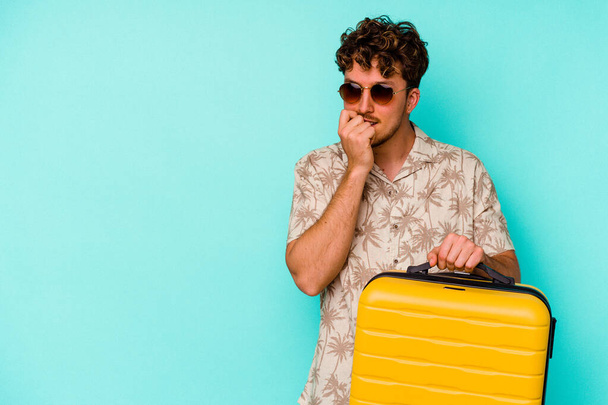 Young traveler man holding a yellow suitcase on blue background relaxed thinking about something looking at a copy space. - Foto, immagini