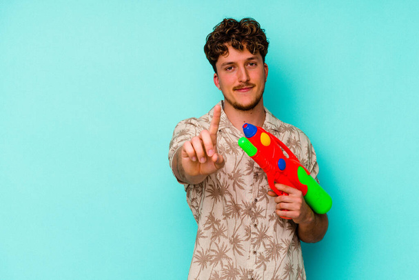 Young caucasian man holding a water gun isolated on blue background showing number one with finger. - Photo, Image