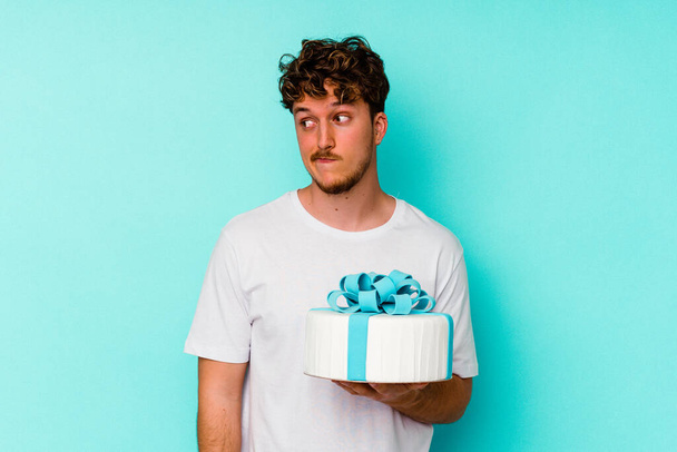 Jonge blanke man houdt een taart geïsoleerd op blauwe achtergrond verward, voelt zich twijfelachtig en onzeker. - Foto, afbeelding