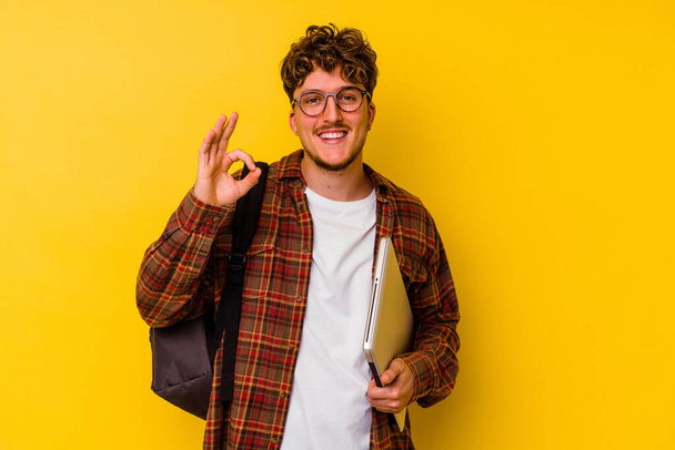 Young student caucasian man holding a laptop isolated on yellow background cheerful and confident showing ok gesture. - Fotografie, Obrázek