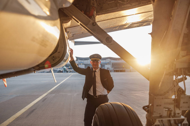 Szakmai férfi pilóta egyenruhában és kalap néz kamera, támaszkodva egy nagy utasszállító repülőgép készen áll az indulásra a repülőtéren naplementekor - Fotó, kép