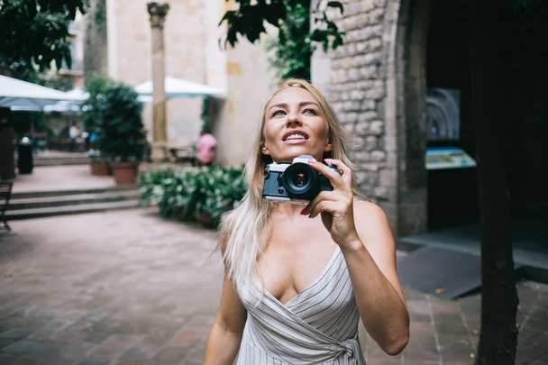 Caucasian woman with retro technology equipment in hand looking up and admire beauty of ancient city enjoying summer international vacations, pretty girl with vintage camera visiting touristic town - Foto, Imagem