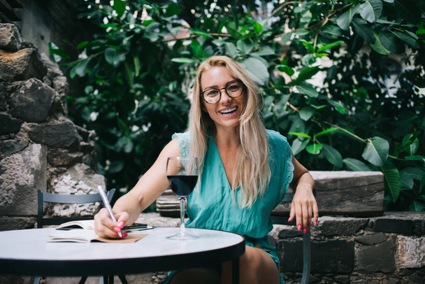 Half length portrait of cheerful female poet in optical eyewear for vision correction smiling at camera during leisure time with wine, happy woman with textbook for planning rejoicing outdoors - Фото, изображение