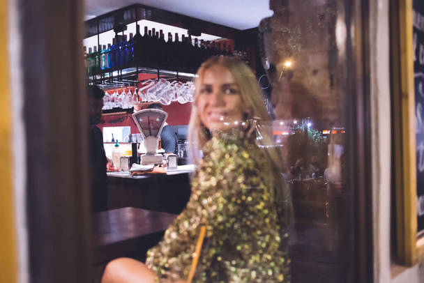 Window view from street inside of disco bar for evening party and funny hang out, Caucasian woman in trendy apparel resting on blurred frontage in public pub, stylish girl enjoying nightlife - Zdjęcie, obraz