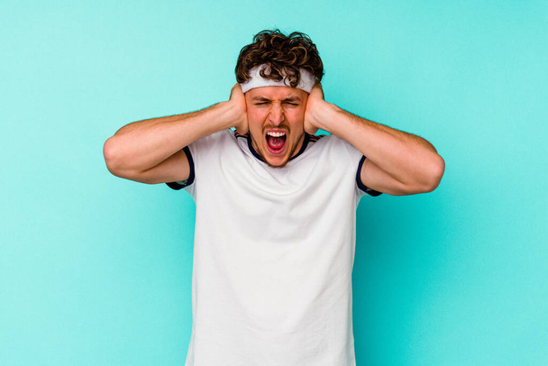 Young sport caucasian man isolated on blue background covering ears with hands trying not to hear too loud sound. - Photo, Image