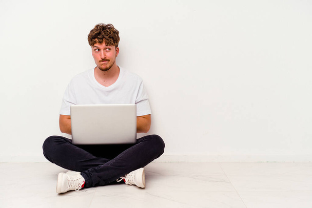 Giovane uomo caucasico seduto sul pavimento tenendo il computer portatile isolato su sfondo bianco confuso, si sente dubbioso e insicuro. - Foto, immagini