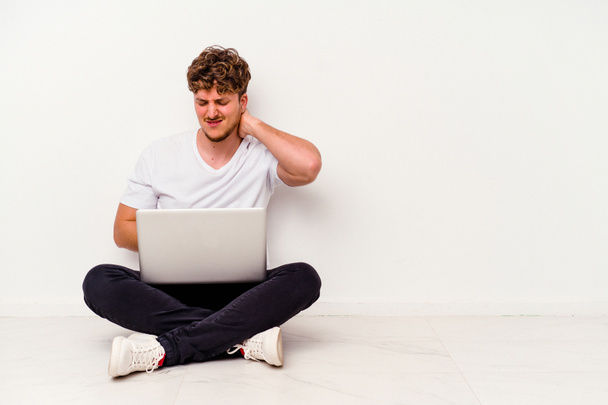 Jonge blanke man die op de vloer zit en een laptop vasthoudt, geïsoleerd op een witte achtergrond, die nekpijn lijdt als gevolg van een zittende levensstijl. - Foto, afbeelding