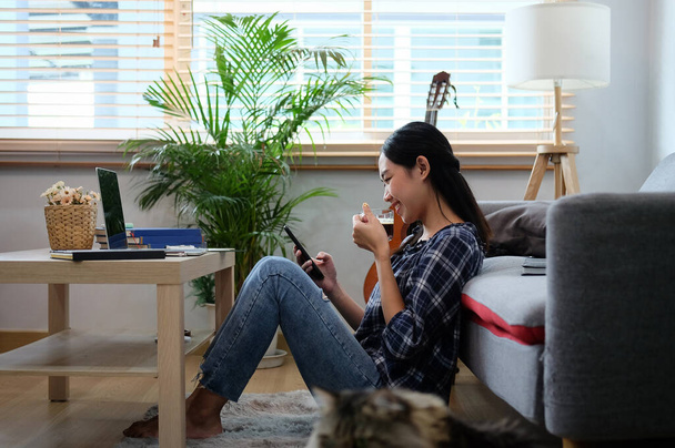 Вид збоку випадкова жінка п'є каву і використовує мобільний телефон у вітальні
. - Фото, зображення