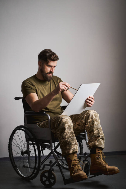 Νεαρός στρατιωτικός που κάθεται σε αναπηρική καρέκλα και ζωγραφίζει σε καμβά - Φωτογραφία, εικόνα
