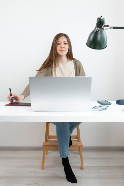 Ładny grafik lub retuszer siedzi przy stole z laptopem i patrzy w ekran. Kobieta w szarym swetrze i niebieskich dżinsach trzymać w ręku ołówek na tablet graficzny. Na biurku telefon i okulary. - Zdjęcie, obraz