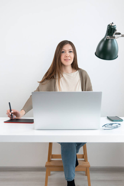 Mooie grafisch ontwerper of retoucher zittend aan tafel met laptop en kijkend in de camera. Meisje in grijs vest en blauwe jeans in de hand potlood voor grafische tablet. Telefoon en bril op het bureau. - Foto, afbeelding