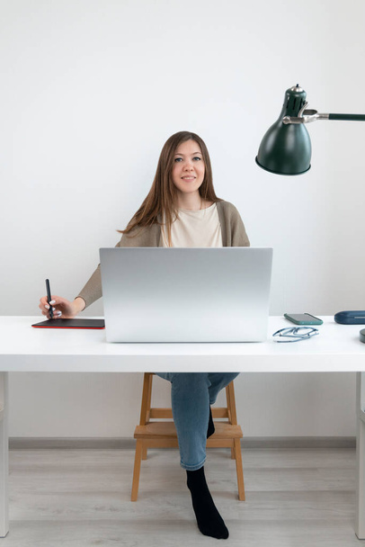 Hezký grafik nebo retuš sedí u stolu s notebookem a dívá se do kamery. Dívka v šedém svetru a modrých džínách drží v ruce tužku na digitální tablet. Bílé pozadí. - Fotografie, Obrázek